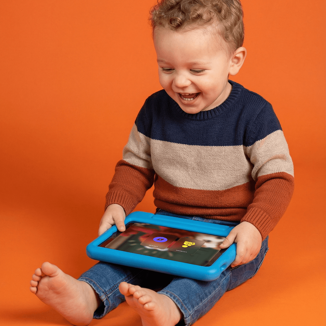 A smiling child in a striped sweater holds a blue tablet while sitting on an orange background.